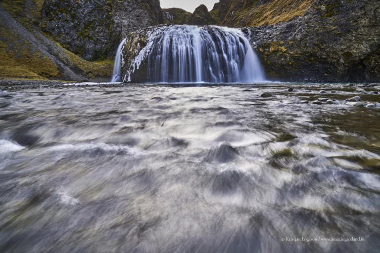 Stjórnarfoss is a small waterfall with an unusually sculpted form: a soft, rounded lip, a wide curtain of water, and a sheltered setting that feels more intimate than the famous “headline” falls along Route 1