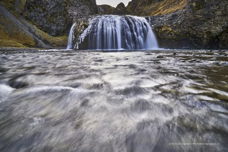 Stjórnarfoss is a small waterfall with an unusually sculpted form: a soft, rounded lip, a wide curtain of water, and a sheltered setting that feels more intimate than the famous “headline” falls along Route 1
