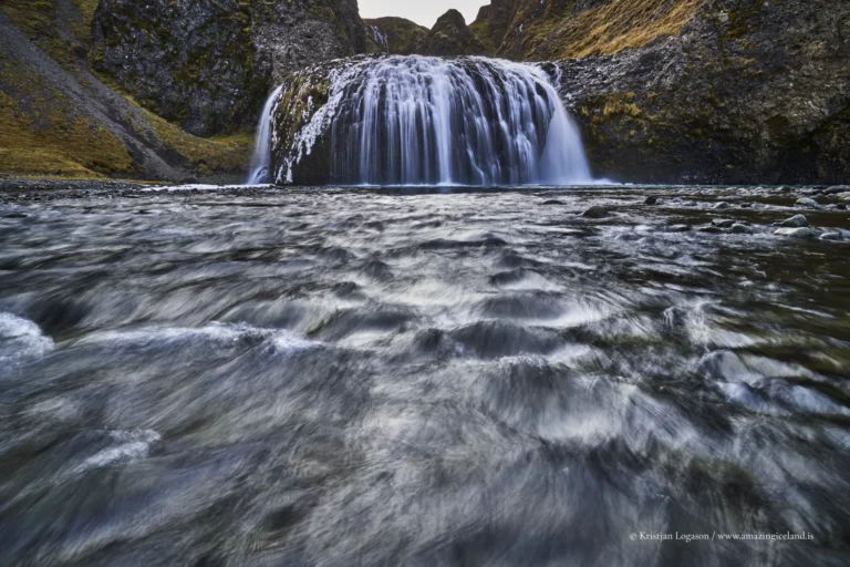 Stjórnarfoss is a small waterfall with an unusually sculpted form: a soft, rounded lip, a wide curtain of water, and a sheltered setting that feels more intimate than the famous “headline” falls along Route 1