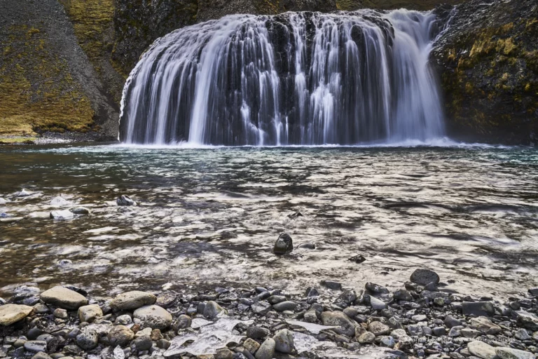 Stjórnarfoss is a small waterfall with an unusually sculpted form: a soft, rounded lip, a wide curtain of water, and a sheltered setting that feels more intimate than the famous “headline” falls along Route 1