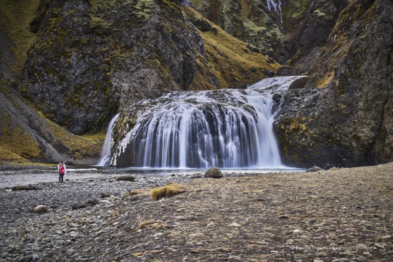 Stjórnarfoss is a small waterfall with an unusually sculpted form: a soft, rounded lip, a wide curtain of water, and a sheltered setting that feels more intimate than the famous “headline” falls along Route 1